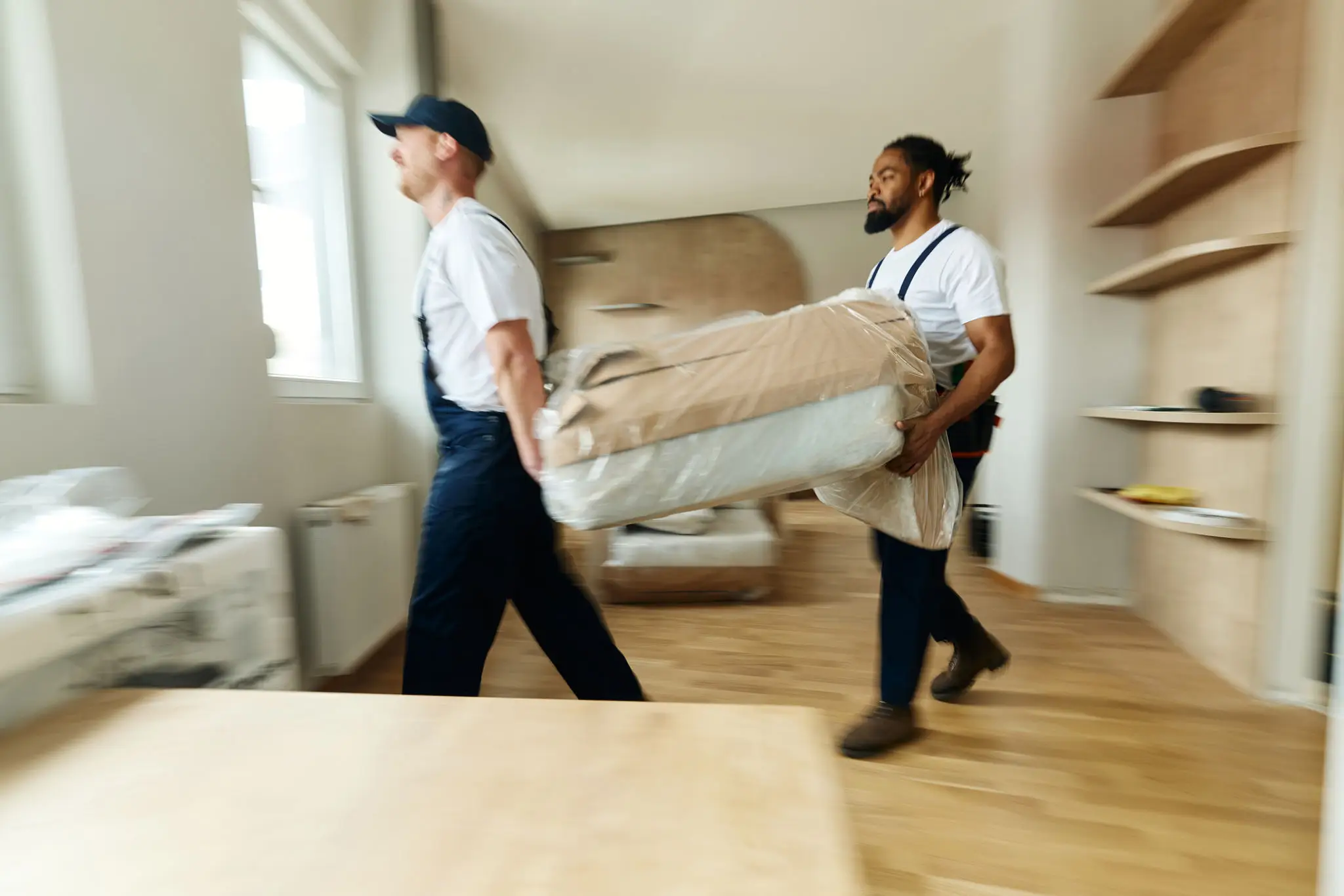 Two movers carrying a large wrapped item through a room.